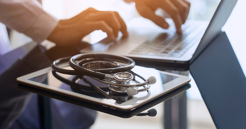 STETHOSCOPE ON IPAD WHILE DOCTOR USES LAPTOP